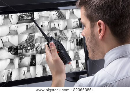 Young Male Security Guard Talking On Walkie-talkie While Monitoring Multiple CCTV Footage On Computer