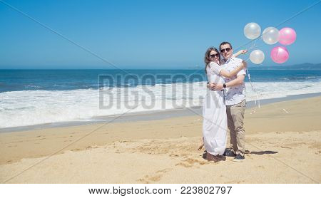 Young romantic couple in white spending vacation or weekend on the beach, celebrating with pink balloons, hugging, for header webside