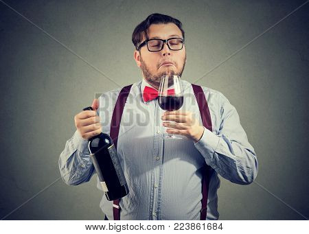 Concentrated man holding bottle of wine and smelling beverage with eyes closed on gray backdrop.