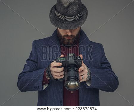 Bearded man in a hat looking down at dslr photo camera. Isolated on grey background.