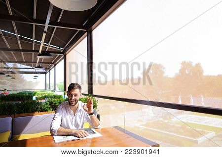 Satisfied magazine editor showing ok geste at cafe table. Personable man with beard wears classic style and looking at camera. Concept of glossy paper edition and writers career.