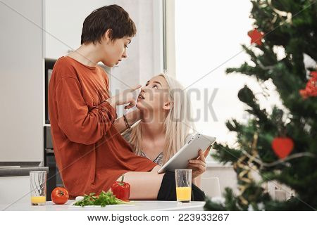 Indoor portrait of young sensual and tender couple of girls, expressing love and attraction while sitting in kitchen and holding tablet on christmas morning. Samesex couple flirt and eat breakfast.