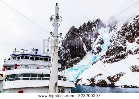 Antarctic Cruise Ship Front View With Mast And Huge Steep Stone Rock Covered With Glacier, Close To 