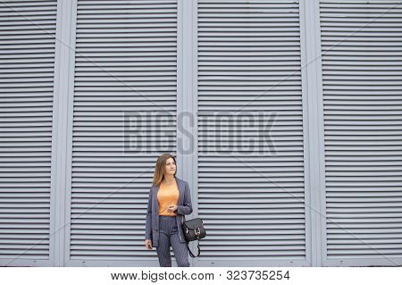 Young Businesswoman In The Morning Stay In The City Against The Background Of The Wall, A Girl In A 