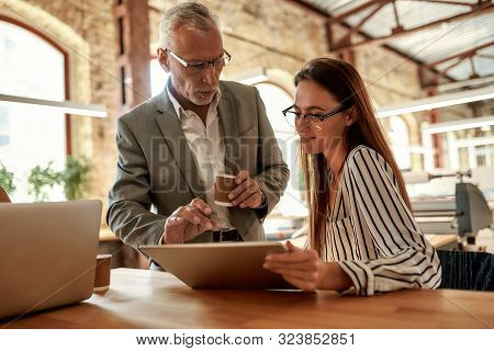 Senior Man In Formal Wear Holding Coffee Cup And Explaining Something To His Young Female Colleague 