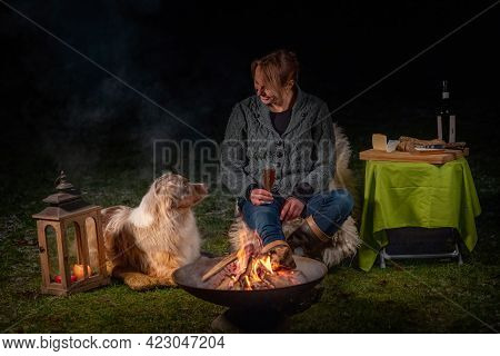 Young Woman Drinks Red Wine By The Campfire In The Forest. Her Australian Shepperd Is Lying Next To 
