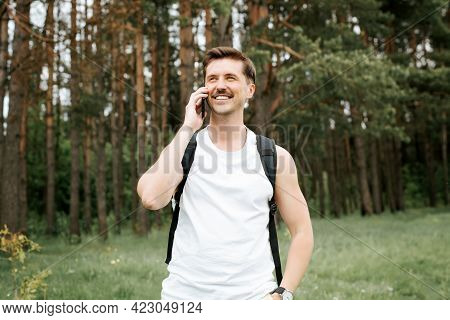 Happy Man Talking On A Cell Phone While Walking In The Woods. Athletic Traveler With Backpack Smilin