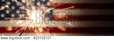 Sparks flying off a burning sparkler in front of the US American flag for patriotic 4th of July celebration.