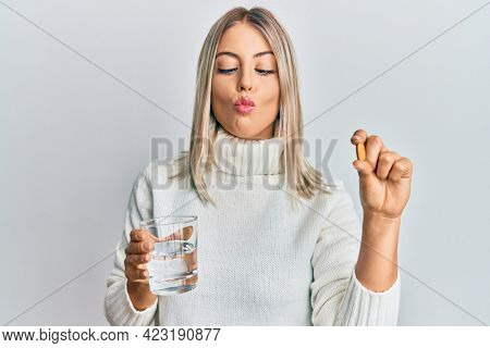 Beautiful blonde woman holding pill and glass of water making fish face with mouth and squinting eyes, crazy and comical. 