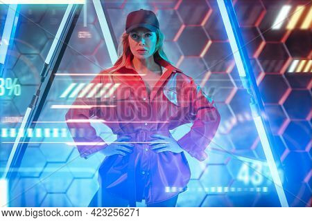 Techno style, disco. Stylish sexy woman in trendy red jacket and baseball cap posing in the light of neon lamps. Fashion shot.