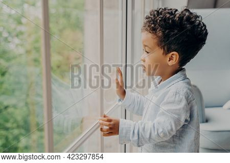 Side View Of Focused Adorable Sweet Kid Boy Looking Outside Through Window With Surprised And Intrig