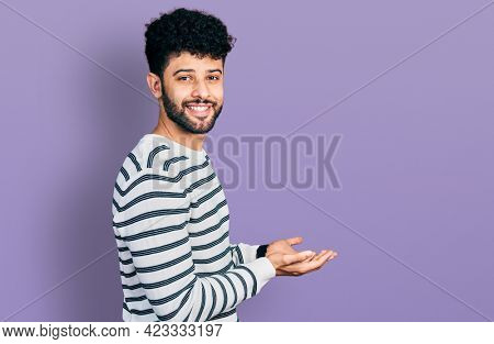 Young arab man with beard wearing casual striped sweater pointing aside with hands open palms showing copy space, presenting advertisement smiling excited happy 