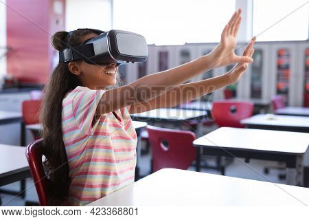 African american girl wearing vr headset gesturing while sitting on her desk in the class at school. school and education concept