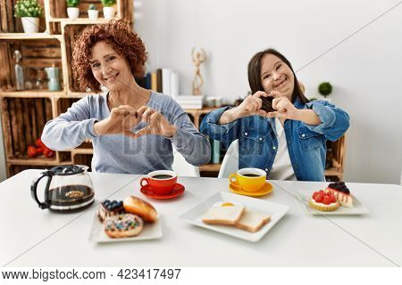 Family of mother and down syndrome daughter sitting at home eating breakfast smiling in love doing heart symbol shape with hands. romantic concept. 
