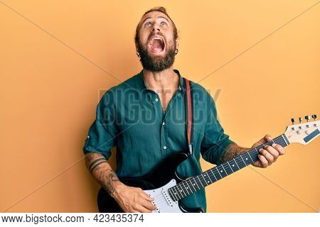 Handsome man with beard and long hair playing electric guitar angry and mad screaming frustrated and furious, shouting with anger looking up. 