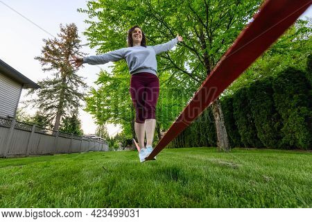 Adventurous White Caucasian Adult Woman Walking On A Slackline Between Trees In A Neighborhood Park.