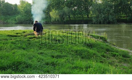 A Fisherman, Leaning Over A Fire On The River Bank, Prepares Fish Soup.