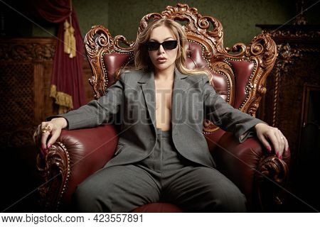 Sexual woman in fashionable pantsuit and black sunglasses alluring in a vintage armchair in a luxury apartments. Wealthy lifestyle. Fashion shot.