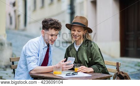 Young Couple Travelers Using Smartphone In City On Holiday, Sitting In Outdoor Cafe.