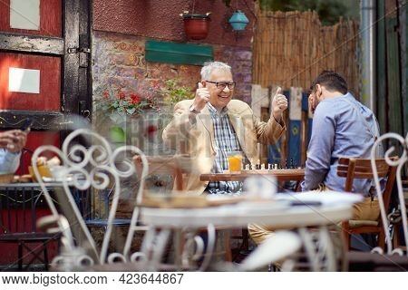 older caucasian male smiling with thumbs up. young guy with his hand over his eyes. sitting in outdoor cafe, playing chess. winning and loosing concept