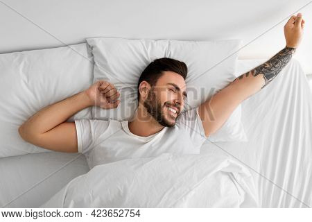 Awakening Concept. Happy Rested Wellslept Man Waking Up In Morning And Stretching Hands, Lying In Be
