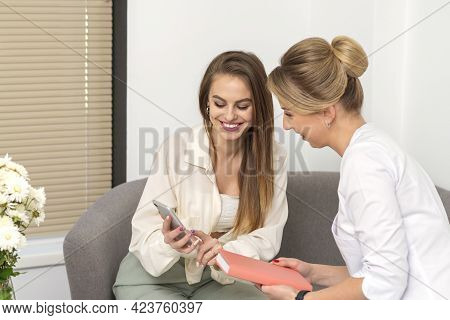Two Young Women Doctor With Pacient Smiling And Looking At Smartphone Sitting In Hospital Office, Co