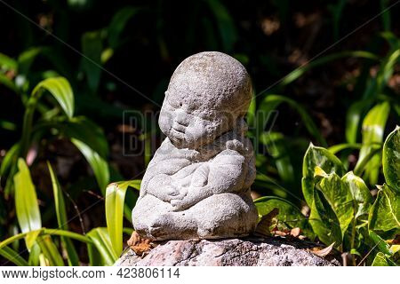 Statue Of Novice Doll In Maditating Act Built From Rough Cement On Rock In Public Garden.
