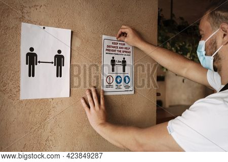 Young white cafe employee in face mask affixing placard while working indoors