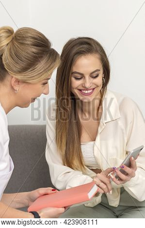 Two Young Women Doctor With Pacient Smiling And Looking At Smartphone Sitting In Hospital Office, Co