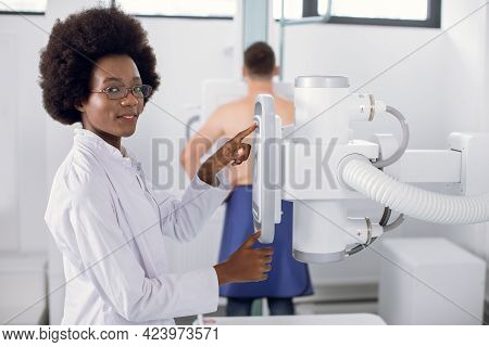Female Aro American Radiologist Technician Setting Up Machine To Perform Chest X-ray On Male Patient