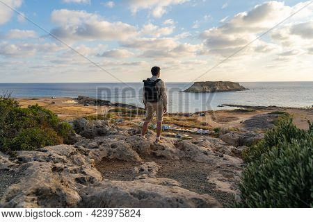 Sea Sunset View. Man With Backpack On Rocks With Beautiful View Of Yeronisos Island Near Coast Of Ag