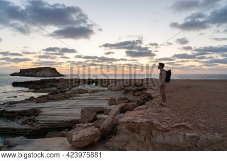 Sea Sunset View. Man With Backpack On Rocks With Beautiful View Of Yeronisos Island Near Coast Of Ag