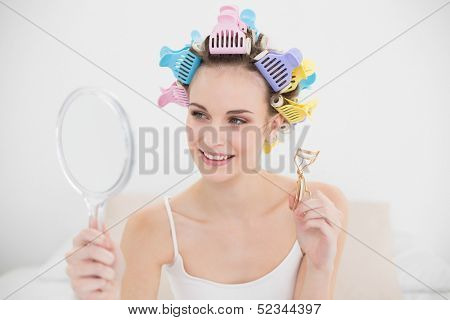 Beautiful natural brown haired woman in hair curlers looking herself in a mirror in bright bedroom