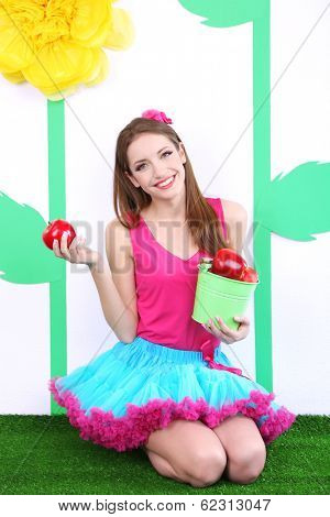 Beautiful young woman in petty skirt holding bucket of apples on decorative background