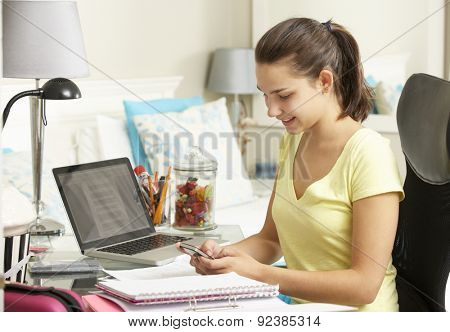 Teenage Girl Studying At Desk In Bedroom Using Mobile Phone