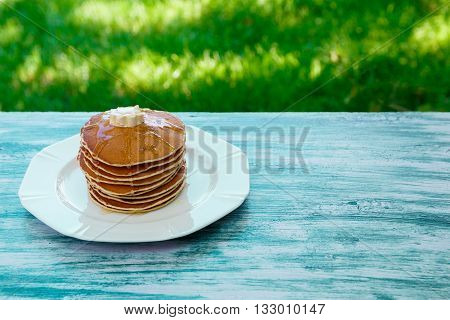 Pancakes with butter and honey on white plate on blue wooden background in garden or on nature background. Stack of wheat golden pancakes or pancake cake closeup.
