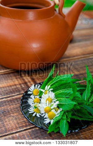 Open brown teapot with flower of chamomile near black plate with fresh mint and chamomile on wooden background. Closeup. Tea concept.