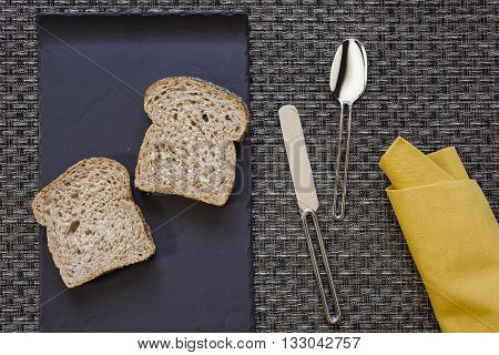 Black flat plate with two slices of bread and mustard yellow folded napkin spoon and knife aside on woven grey thick wire warp textured background