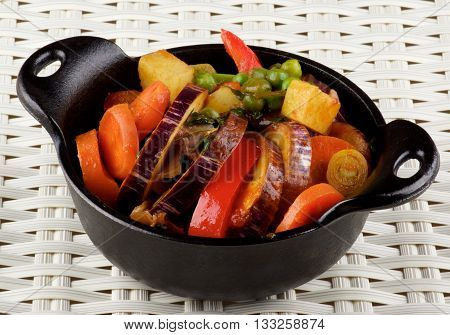 Delicious Homemade Colorful Vegetables Ragout with Striped Eggplant Carrots Potatoes Leek Red Bell Pepper and Green Pea in Black Iron Stewpot closeup on Wicker background