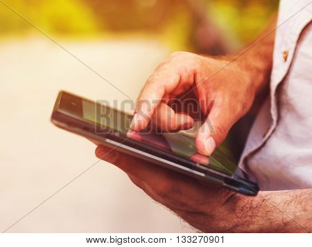 Smart casual man with digital tablet outdoors, close up. Focus on finger. Sunkissed effect with sunflares. Modern remote work concept.