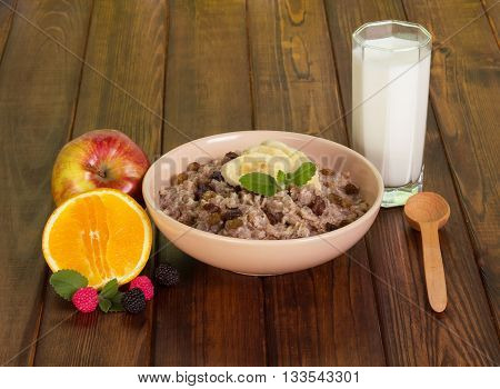A bowl of oatmeal with raisins, fruit, a glass of milk and a wooden spoon on dark wooden background.