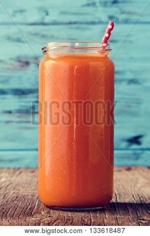 closeup of an orange detox smoothie served in a glass jar with a red drinking straw patterned with white dots, on a rustic wooden surface