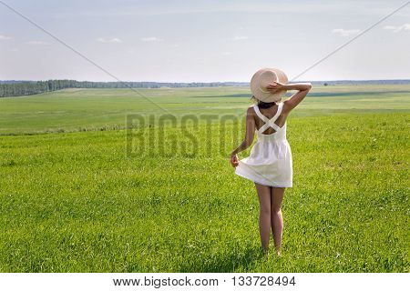 young girl with long dark hair standing on a green field with green grass backs to the camera in a short white summer dress and straw hat and then removes it. throws up his hands and enjoying nature. in the right corner of the screen