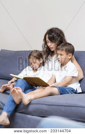 Family Concepts. Mother With Two Children Reading A Book Together On Couch Indoors.vertical Image