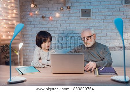 Grandfather And Grandson Are Watching Video At Table At Night At Home.