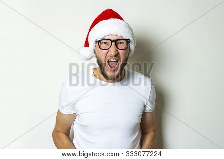 Young Man In Glasses And A Santa Claus Hat Shouts On A White Background. Christmas Concept. Gesture 