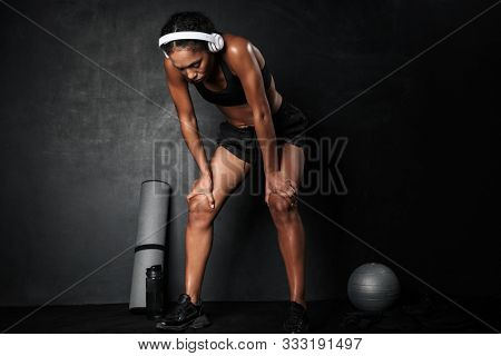 Confident young african fitness woman resting after workout with gym equipment isolated over black background, listening to music with wireless headphones