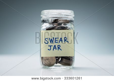 Close-up Of A Glass Jar With Swear Jar Text Filled With Coins Against Grey Background