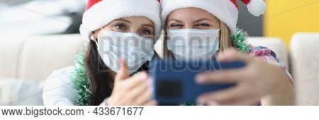 Two Women In Protective Medical Masks And Santa Claus Hats Take Selfie On Smartphone