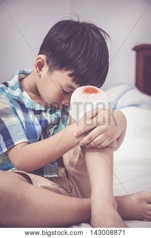 Childhood accidents. Sadness asian boy worry about wound on his knee with bandage. Child injured on bed in bedroom. Human health care and medicine concept. Vintage tone effect.